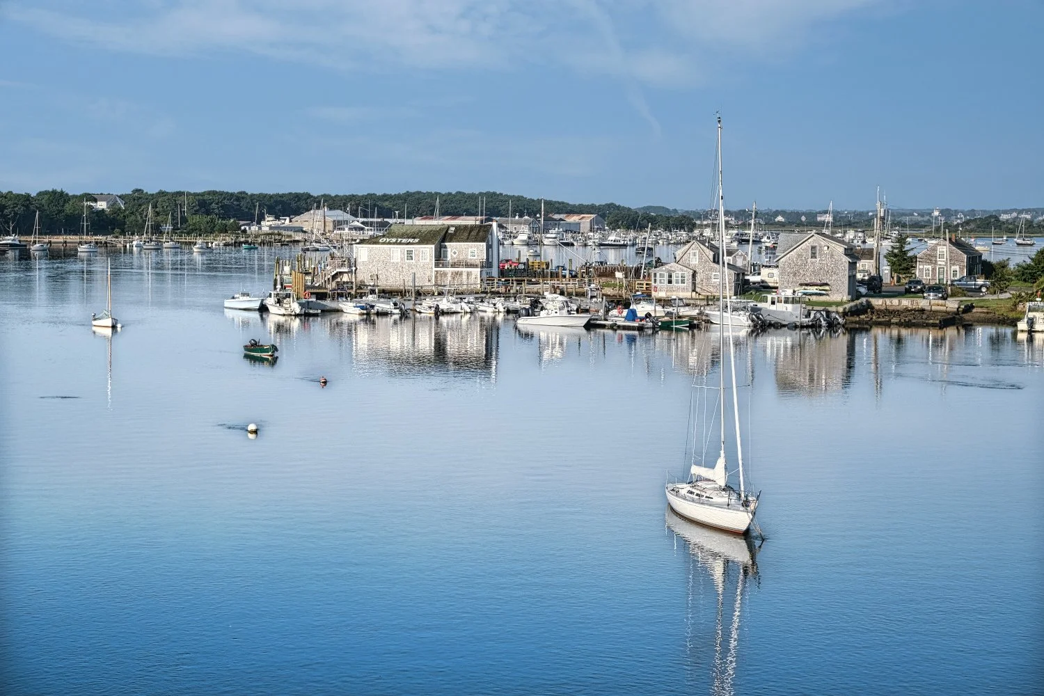 A peaceful harbor scene featuring a calm body of water with several small boats and a sailboat with a tall mast. In the background, there are wooden buildings, some labeled 'OYSTERS,' and a cluster of houses and sailboats along the shoreline under a 