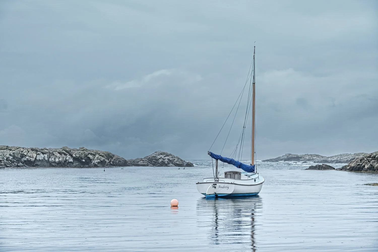 A small sailboat named Audrey floating on calm water with rocky islands in the background under a cloudy sky.