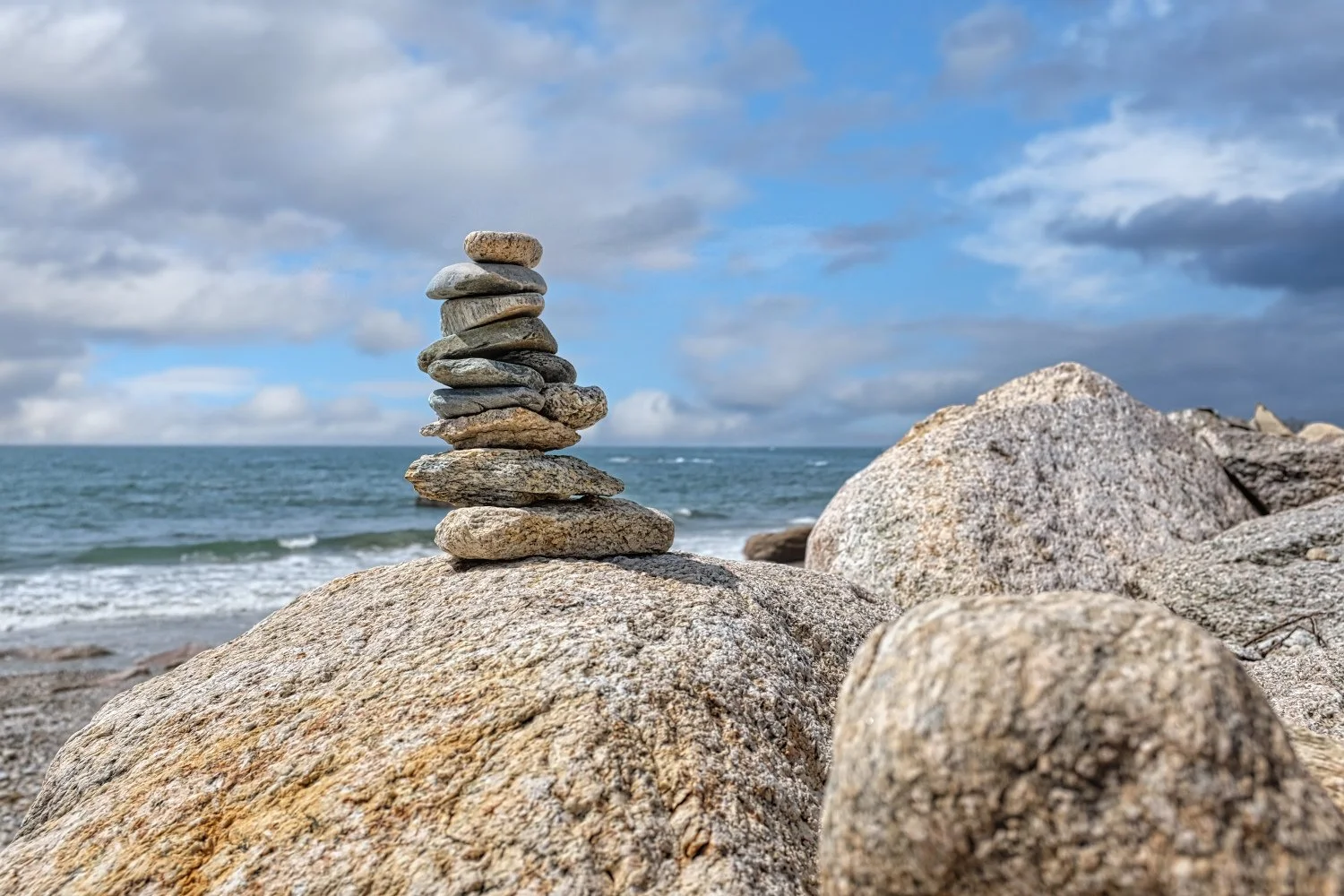 Stacked rocks on a large boulder at the beach with ocean waves and cloudy sky in the background.