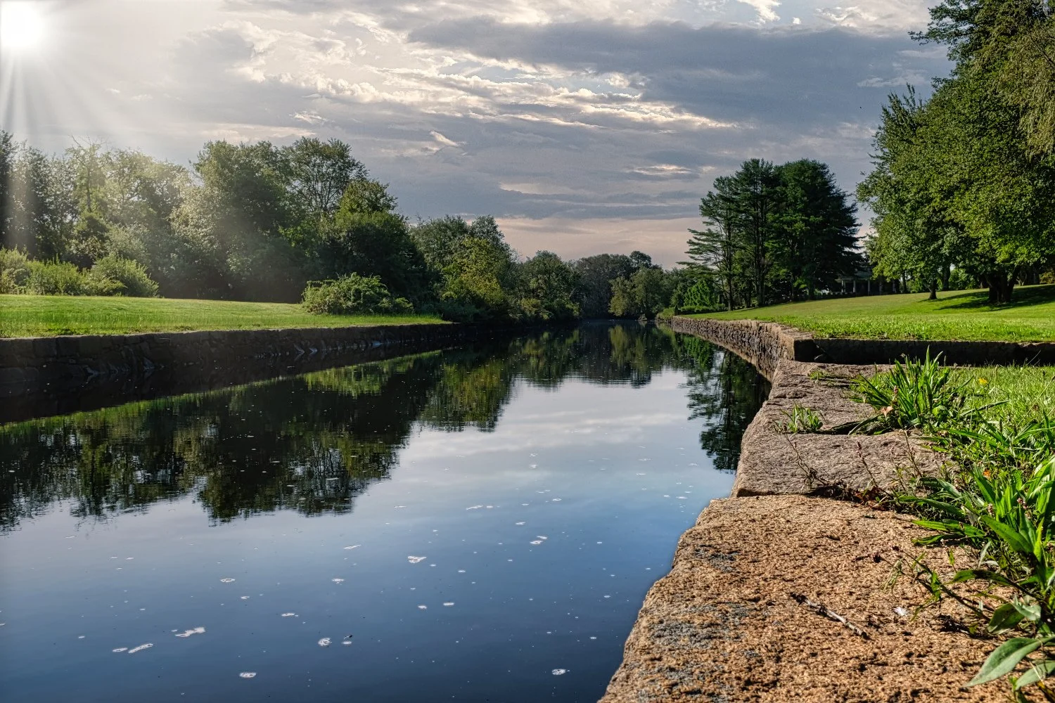 A calm river with a stone edge, surrounded by green trees and grass, under a partly cloudy sky with sunlight.