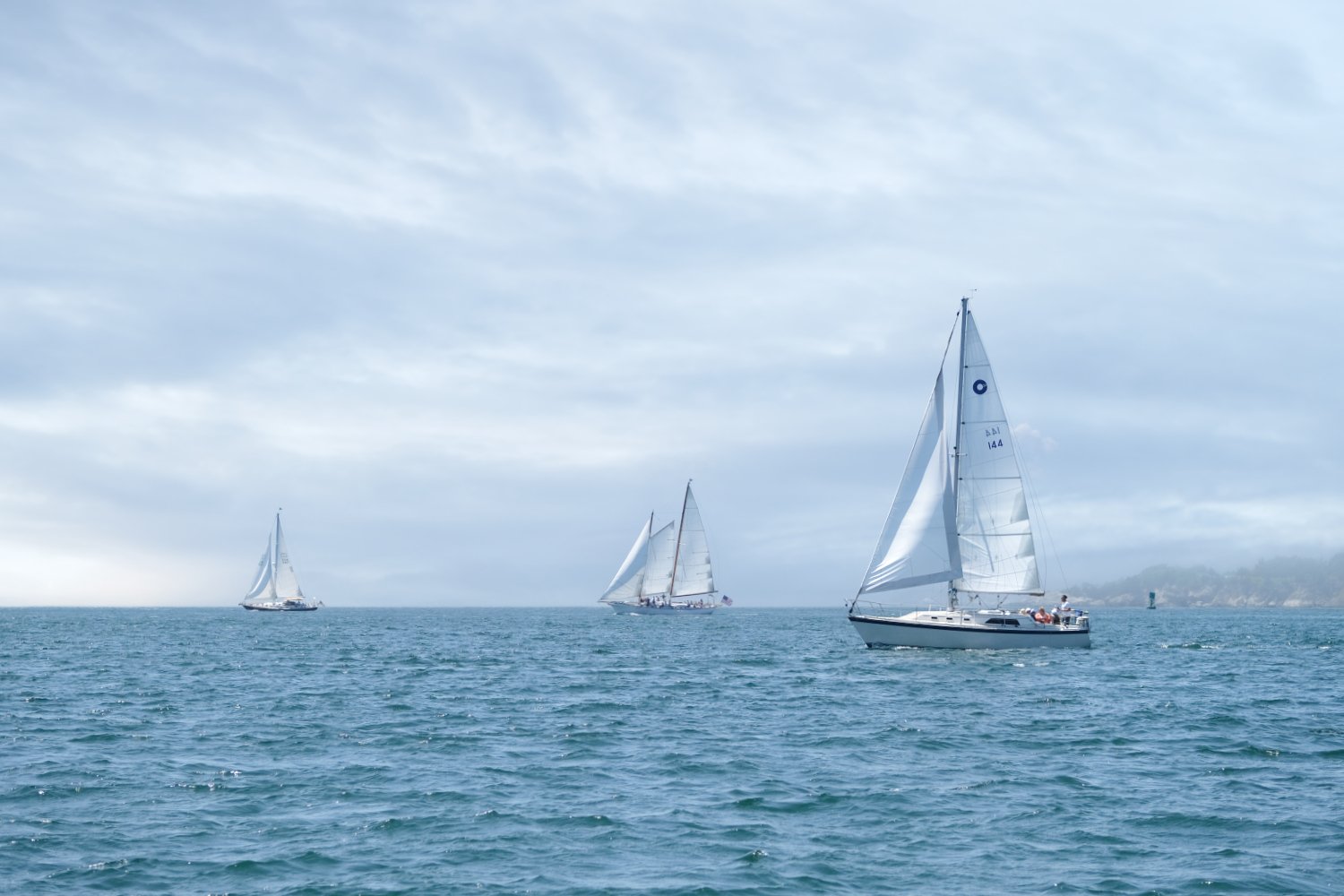 Three sailboats on the ocean with cloudy sky and distant land in the background.