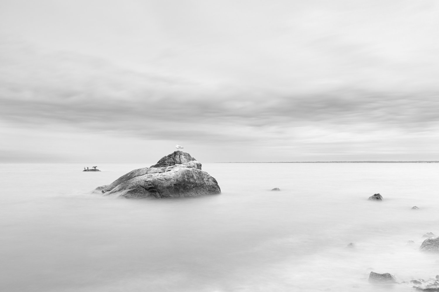 Black and white photo of rocks in calm water near the shore under a cloudy sky.