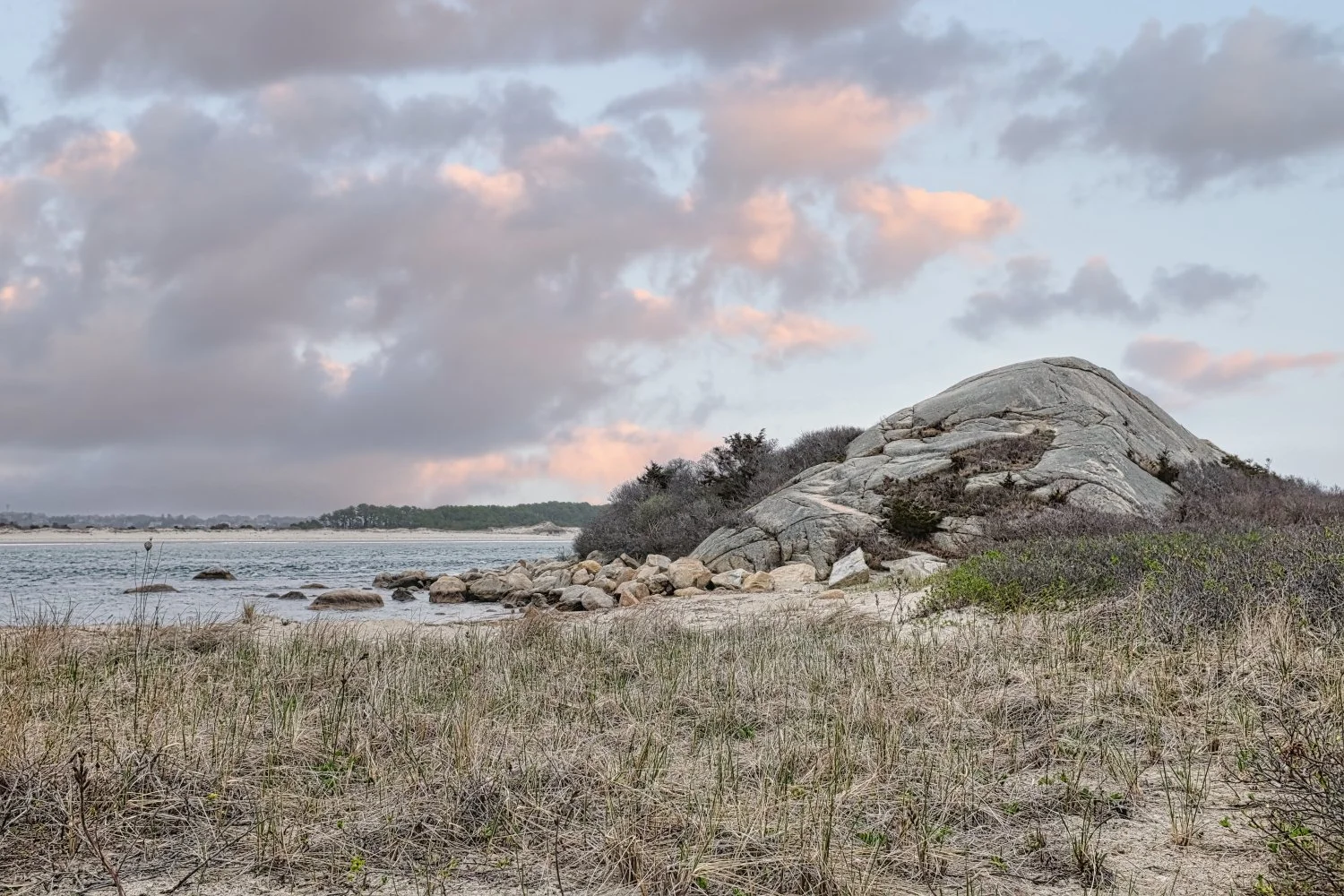 A natural landscape featuring a rocky hillside beside a beach with sand and sparse grass. A calm body of water with small rocks along the shoreline and a distant landmass with trees under a cloudy sky at sunset or sunrise.