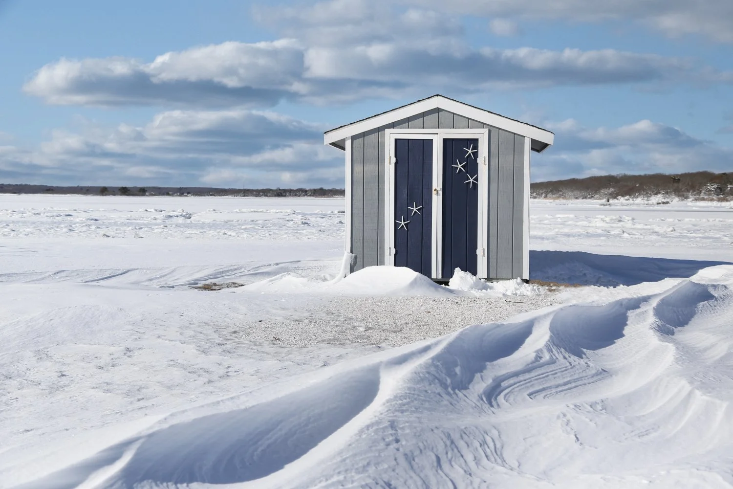 A small, gray and white beach hut with starfish decorations on the door, located in a snow-covered landscape during daytime with a partly cloudy sky.
