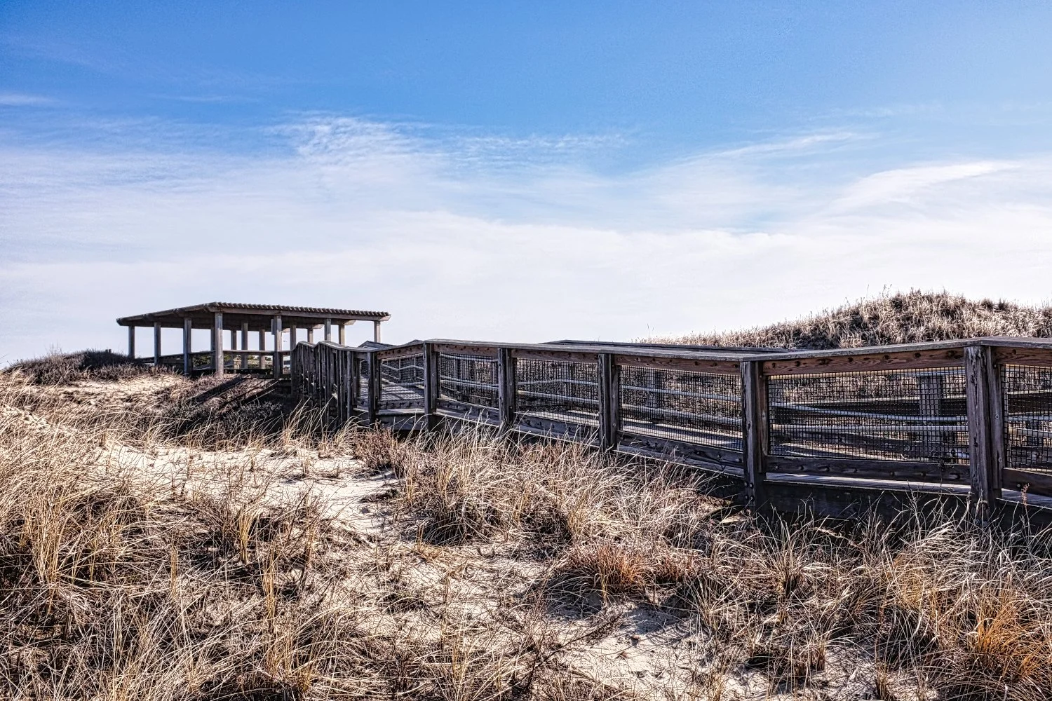 Wooden walkway leading to a shaded pavilion on sand dunes covered with dry grass under a blue sky with scattered clouds.