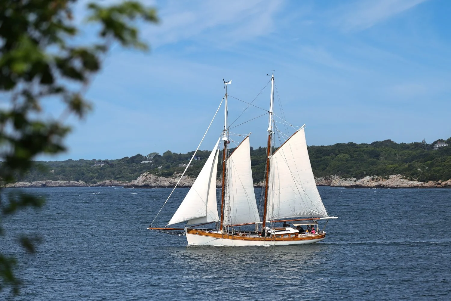 A vintage wooden sailboat with white sails on calm blue water, with a lush green shoreline and hills in the background under a partly cloudy sky.