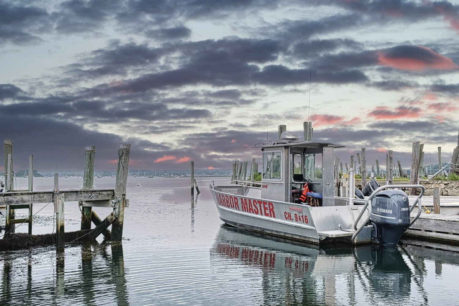 A small boat named 'Harbor Master' docked at a wooden pier during sunset, with a cloudy sky and calm water in the background.