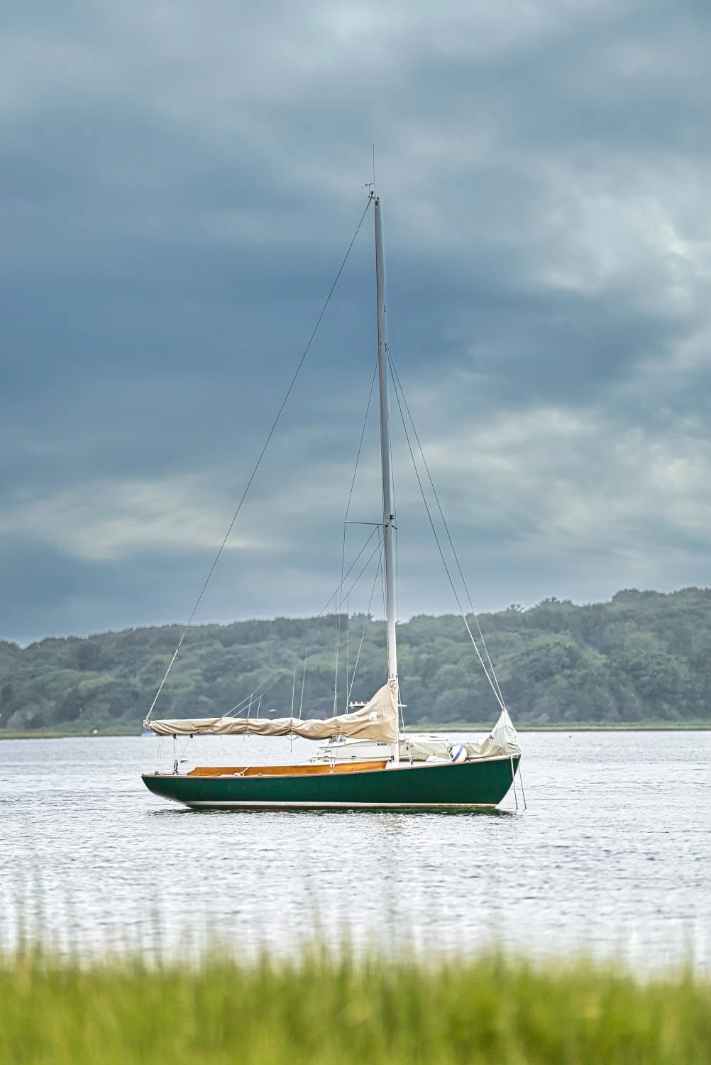 A sailboat floating on calm water with a cloudy sky and lush green hills in the background.