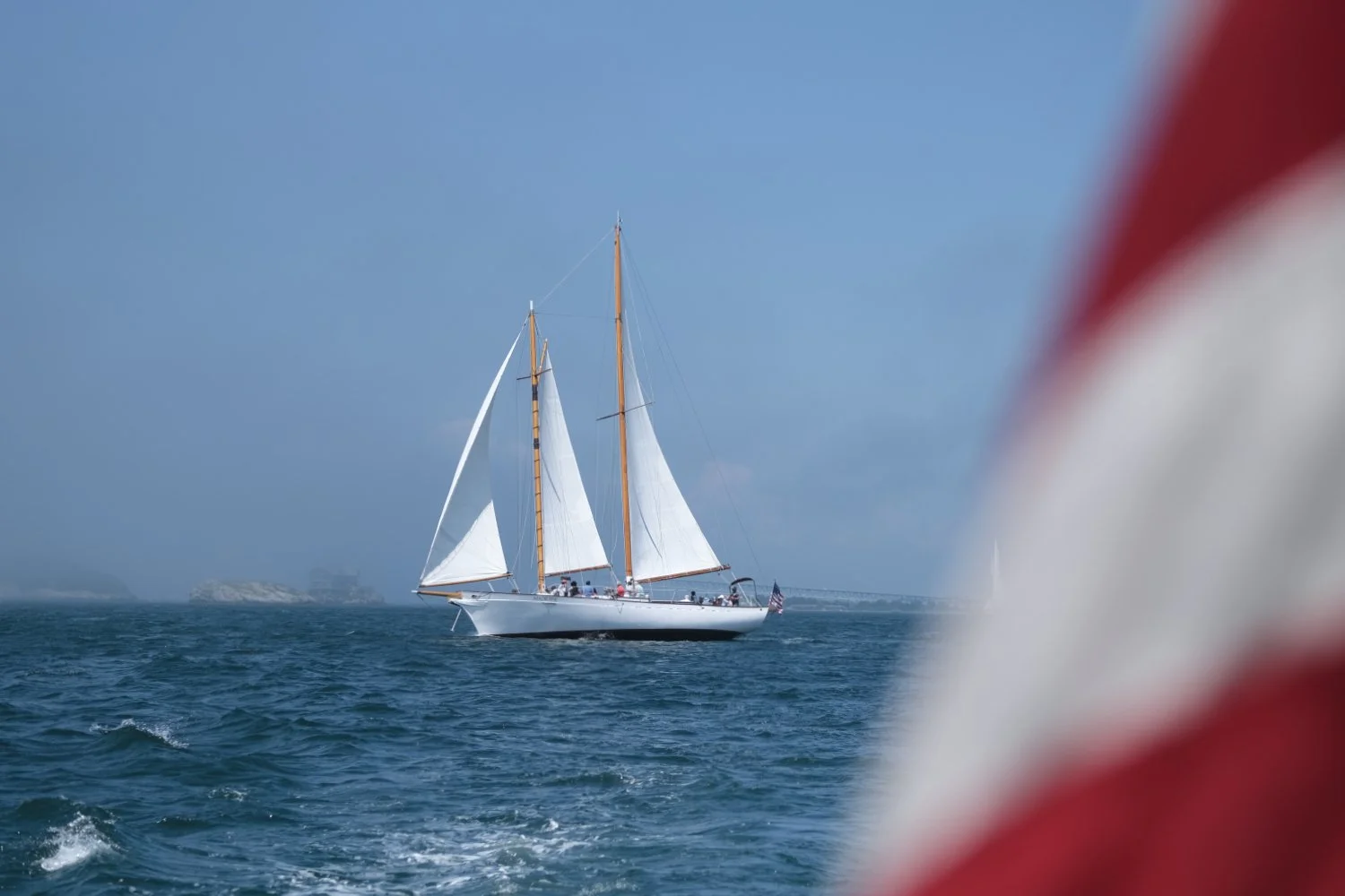 A sailboat on calm blue water with a cloudy sky in the background, partially obscured by a red and white buoy in the foreground.