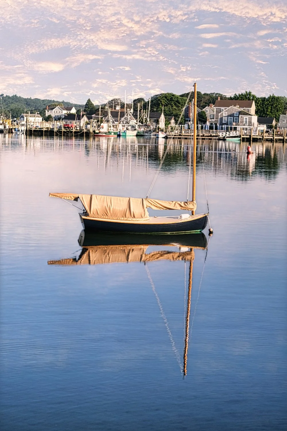 A sailboat with a beige cover on a calm body of water, reflecting the boat and a village with houses and trees in the background under a partly cloudy sky at sunset.