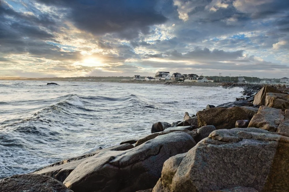 Coastal scene with large rocks in the foreground, ocean waves, and houses along the shore under a cloudy sky at sunset.