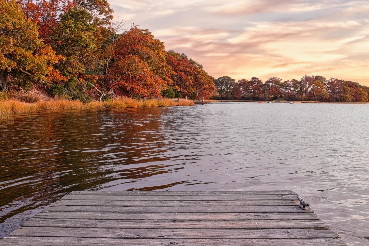 A lakeside scene during autumn with colorful trees along the shoreline, a wooden dock extending into calm water, and boats floating in the distance under a partly cloudy sky at sunset.