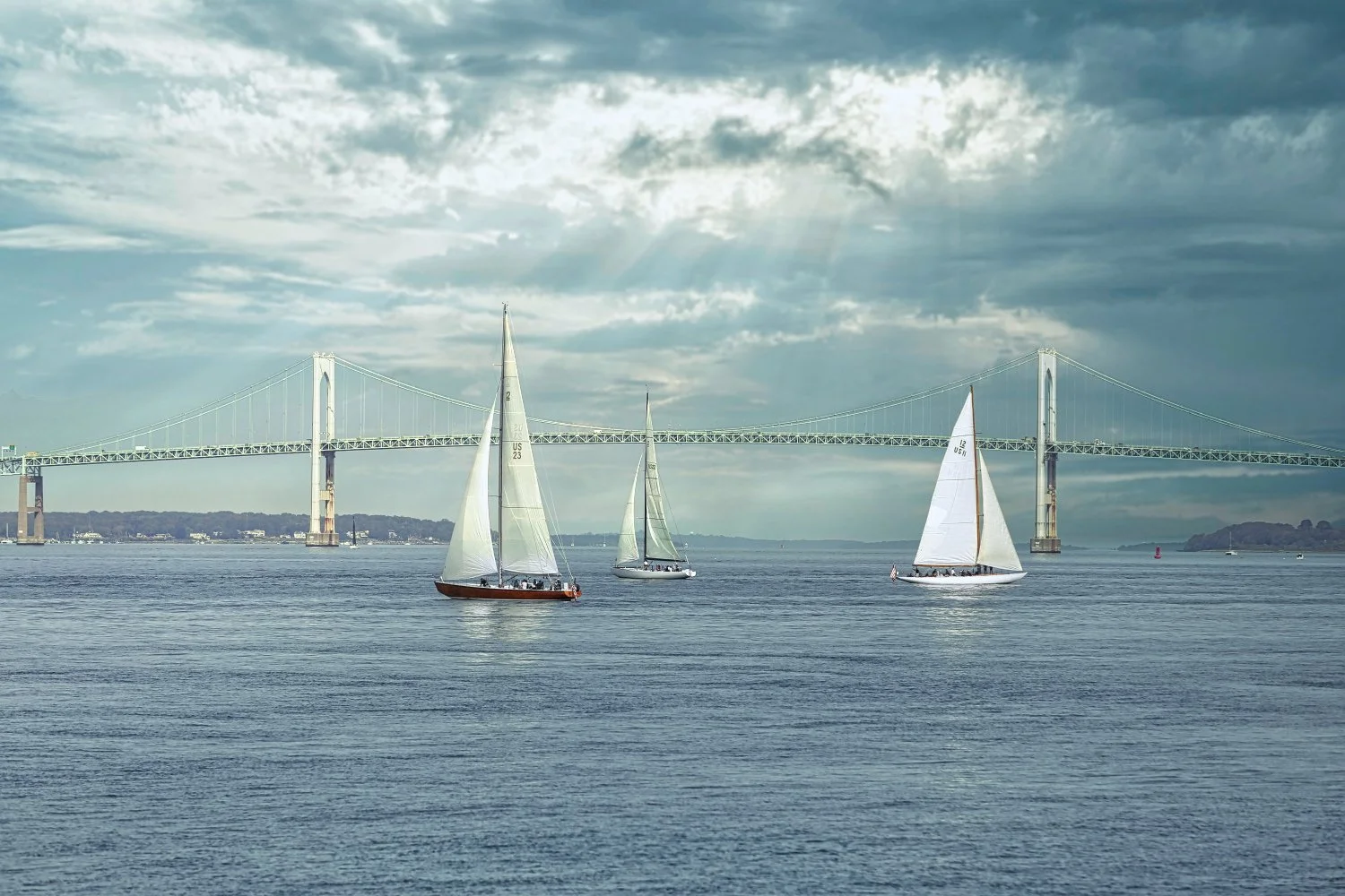 Three sailboats on the water with a large bridge in the background under a cloudy sky.