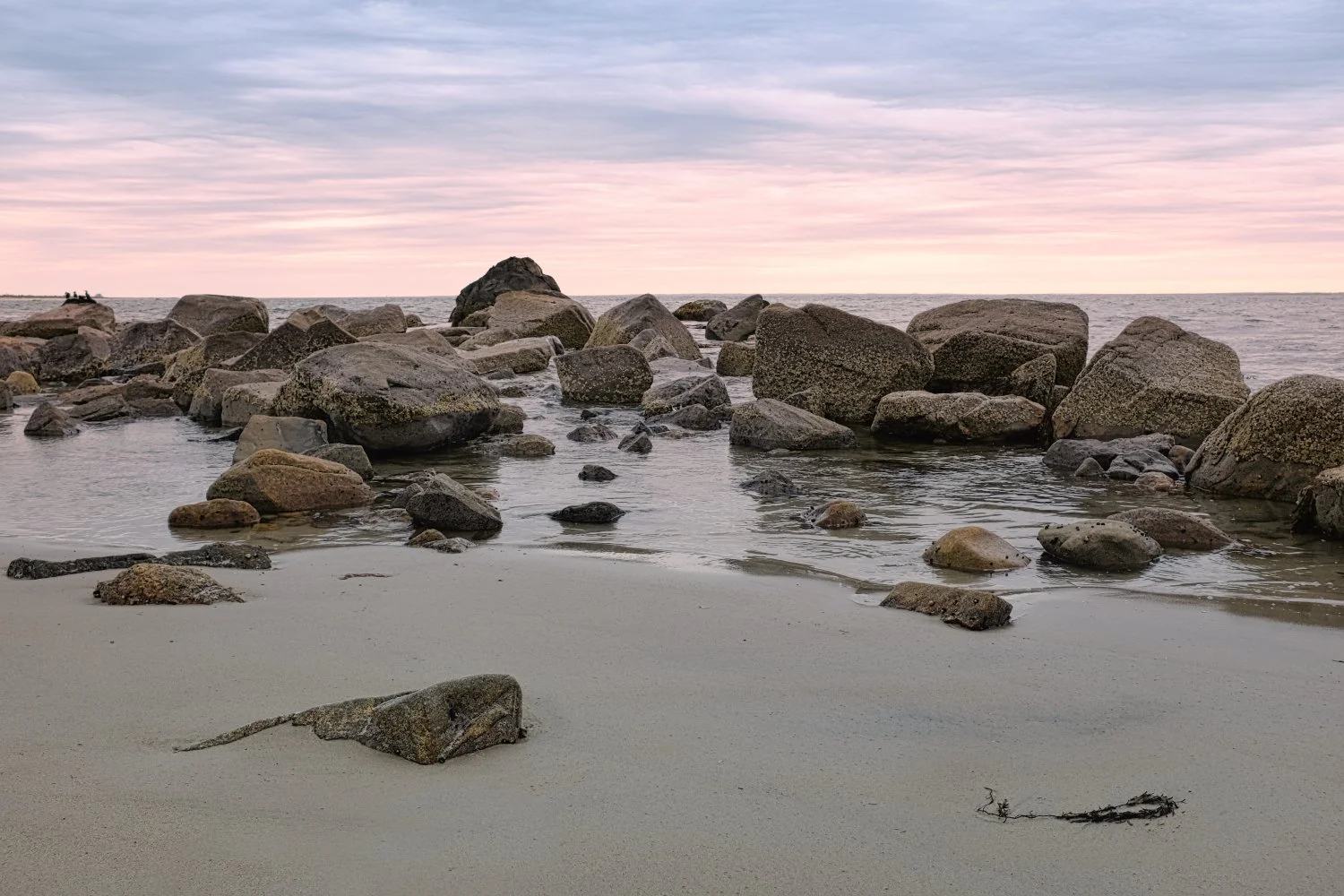A beach scene at sunset with large rocks in the water and a pastel-colored sky.