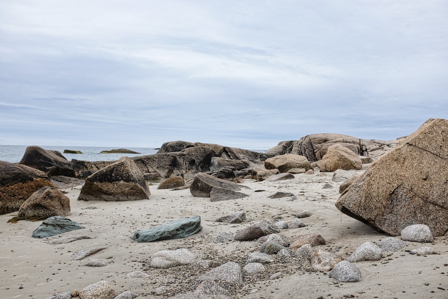A rocky beach with large boulders and smaller stones scattered across the sand, with the ocean in the background under a cloudy sky.