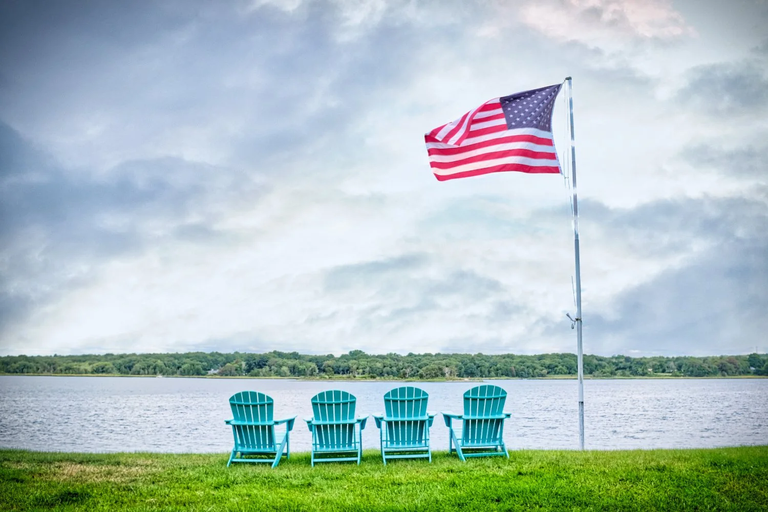Four turquoise Adirondack chairs facing a lake, with an American flag flying on a flagpole nearby, under a cloudy sky.