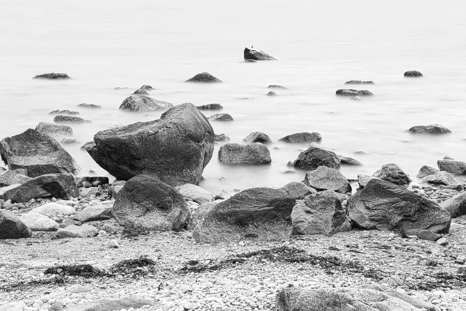 Black and white photo of a rocky shoreline with large rocks and pebbles, and calm water extending into the distance.