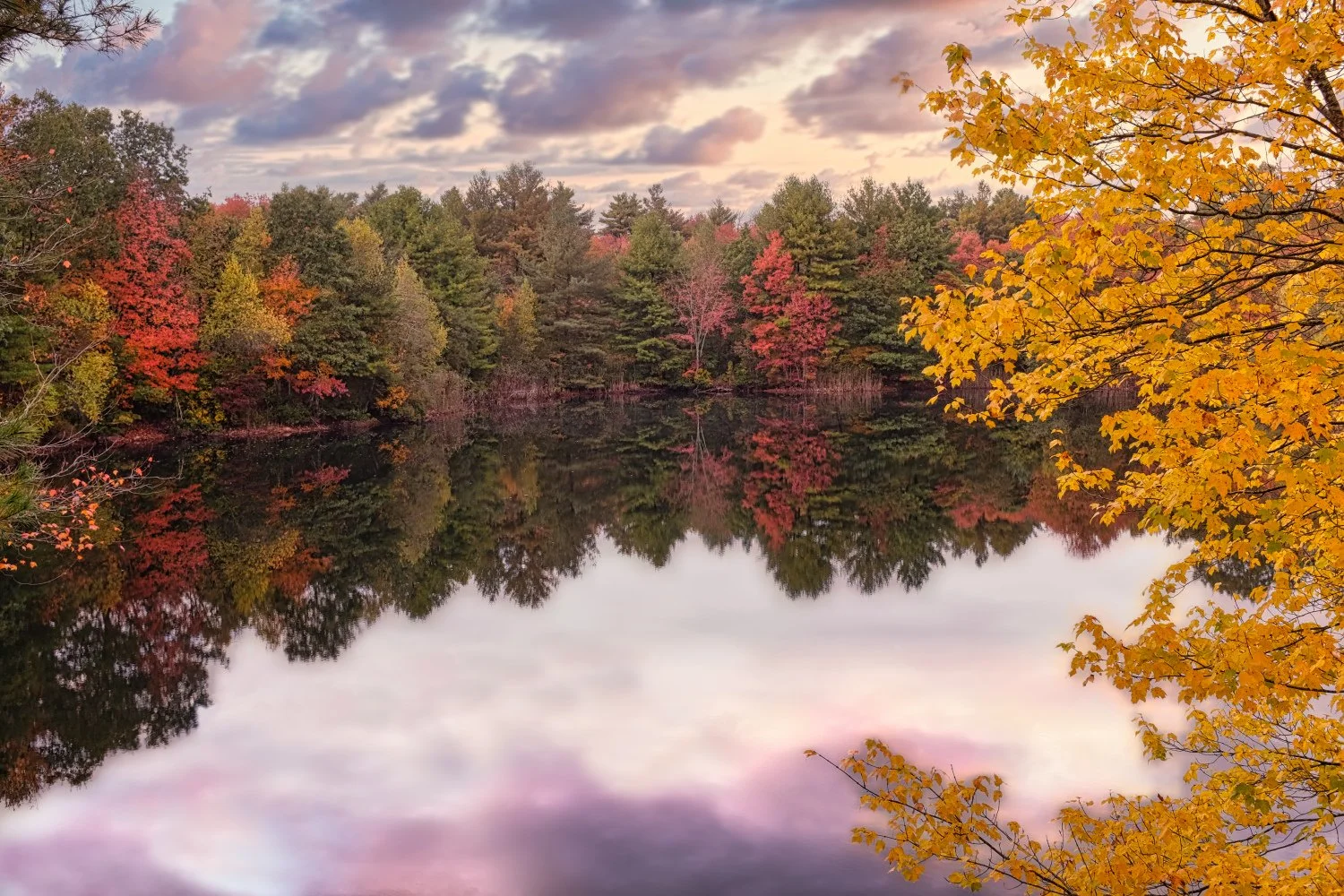 A peaceful lake surrounded by trees with vibrant fall foliage in shades of red, orange, yellow, and green, reflecting on the calm water under a partly cloudy sky at sunset.