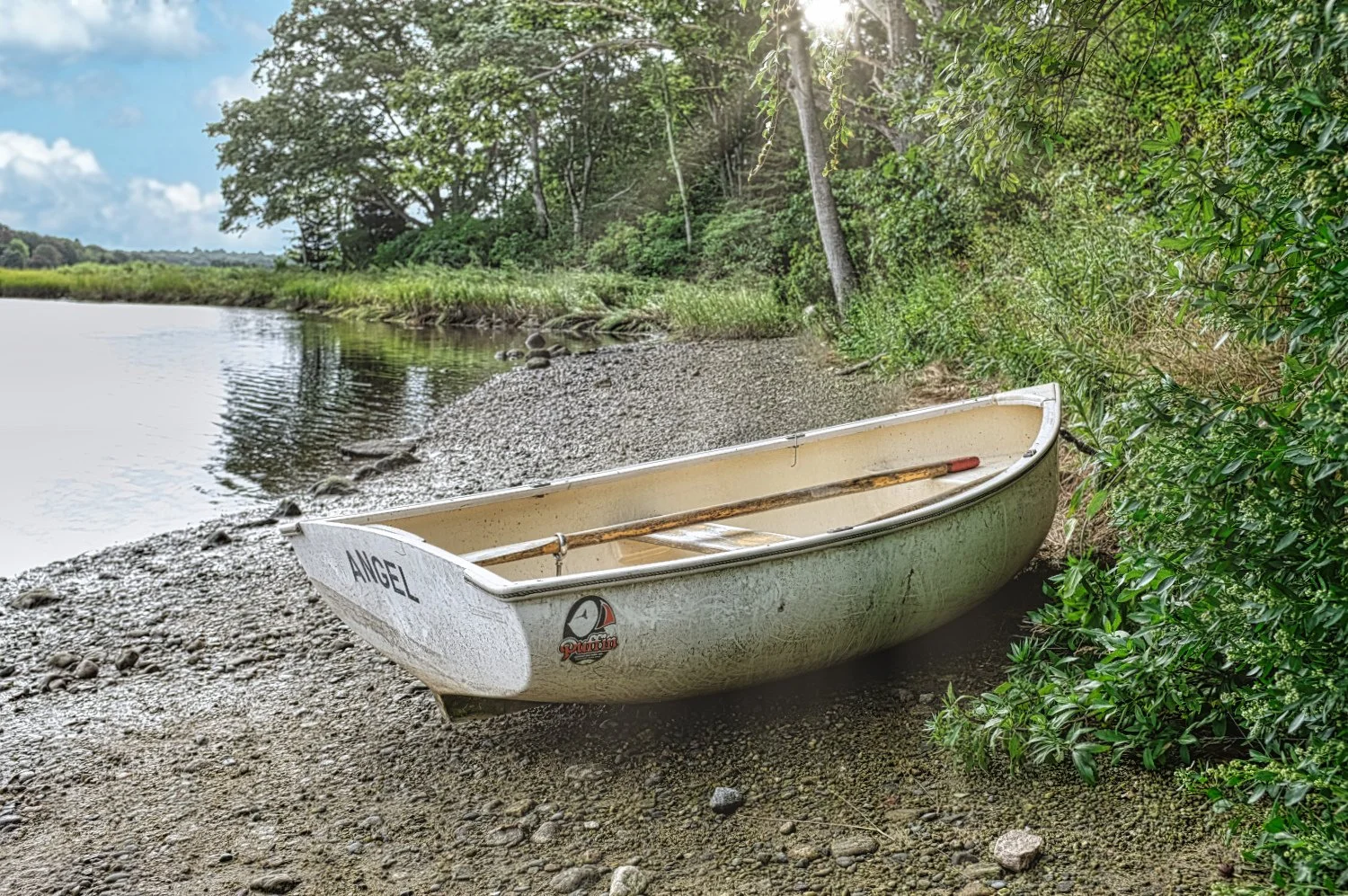 A white rowboat named 'Angel' resting on a pebbly shoreline next to green bushes and trees by a calm river with a cloudy sky above.