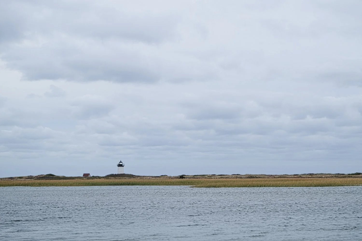 A body of water with a strip of land in the background featuring a lighthouse and a small red-roofed building under a cloudy sky.