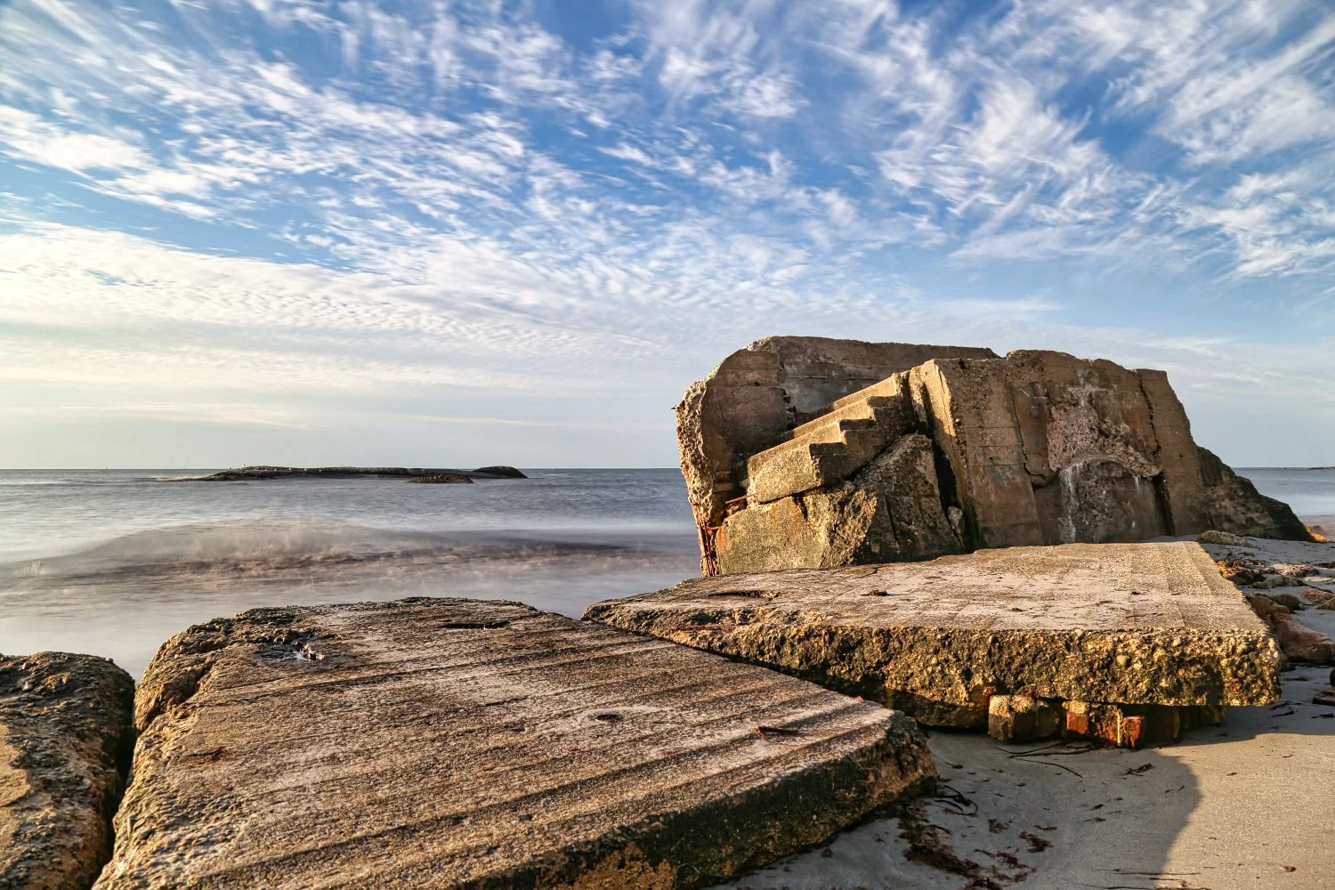 Large weathered concrete blocks and ruins on a sandy beach with the sea and a partly cloudy sky in the background.