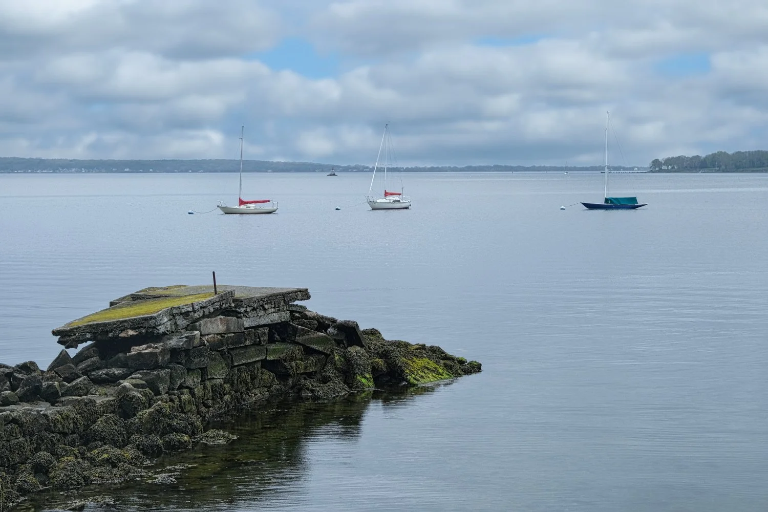 Three sailboats floating on calm water near a stone jetty under a cloudy sky.