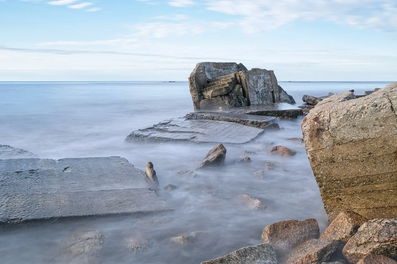 Long exposure photo of ocean waves washing over rocks and concrete slabs along the shoreline under a partly cloudy sky.
