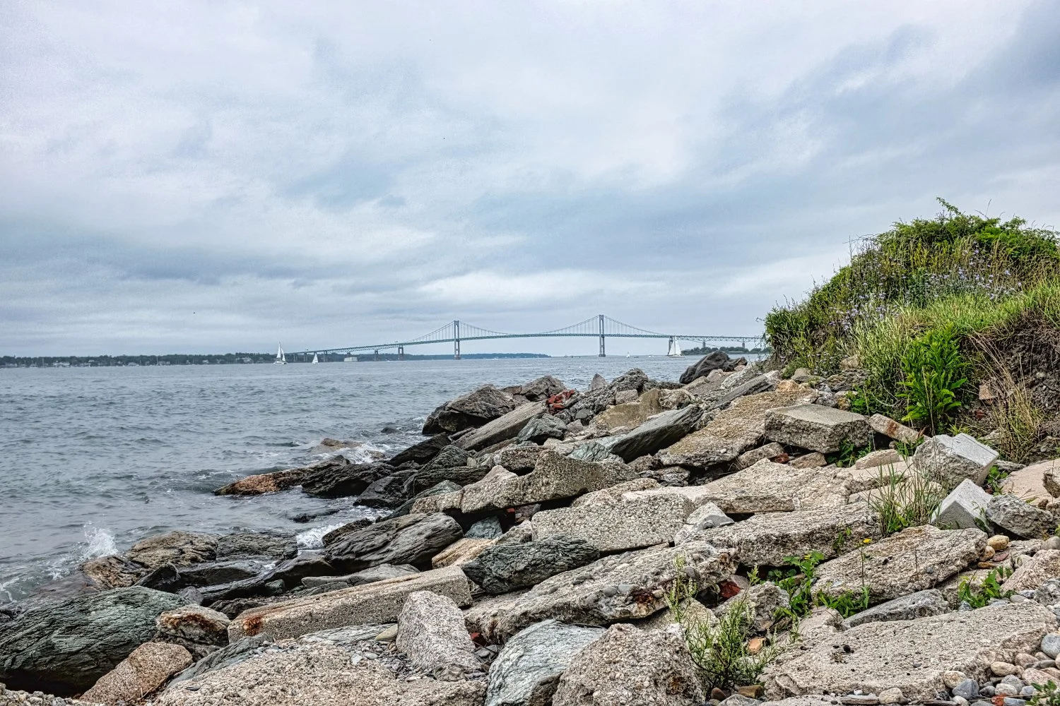 View of a rocky shoreline with a bridge in the distance under an overcast sky