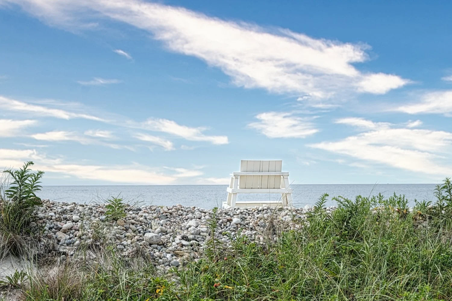 A white bench on a pebble beach facing the ocean under a partly cloudy sky with green plants in the foreground.
