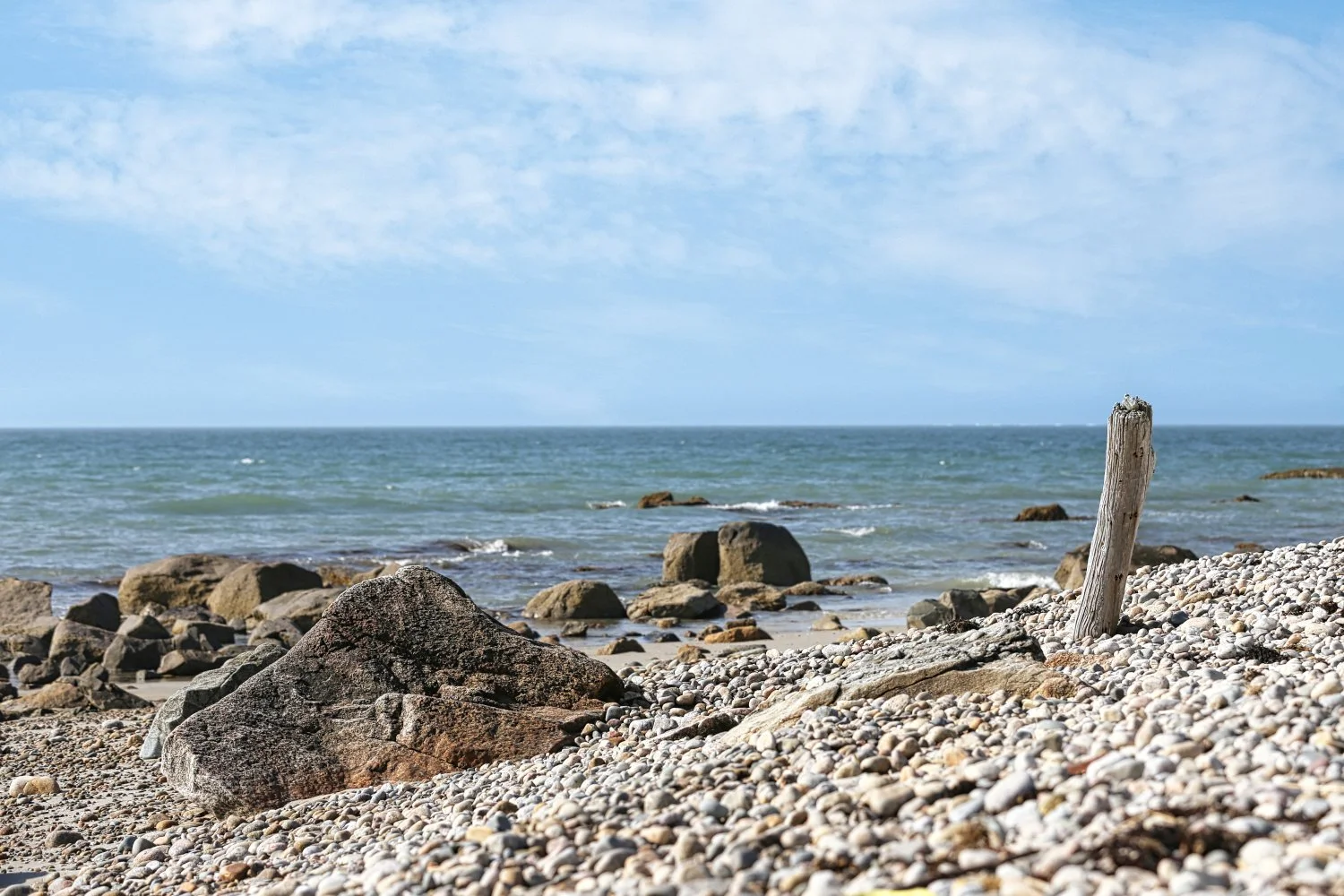 A rocky beach with an ocean view under a partly cloudy sky, featuring a weathered wooden post among rocks and pebbles.