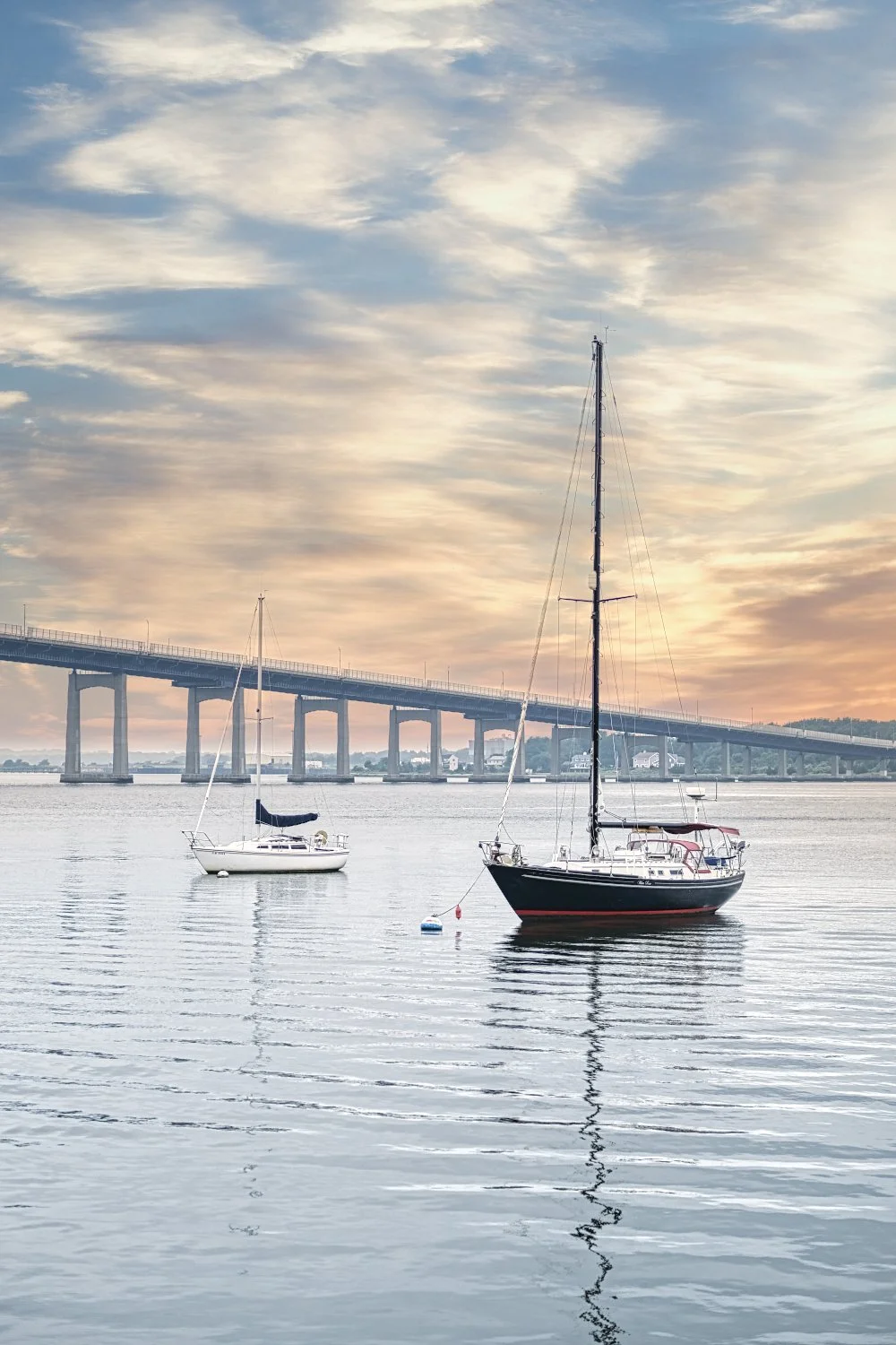 Two sailboats anchored on calm water near a bridge during sunset, with a partly cloudy sky above.