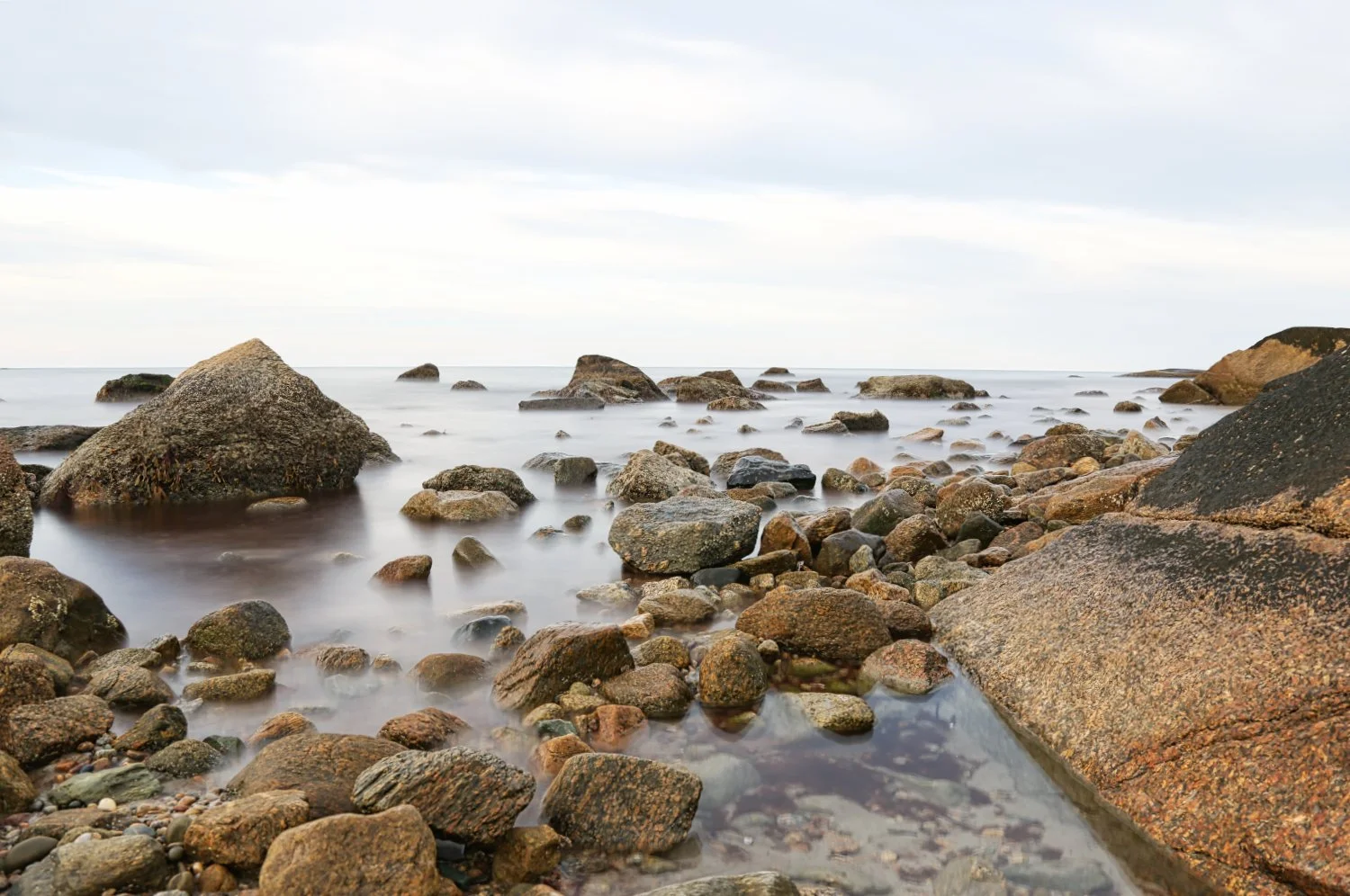 Rocky shoreline with large boulders and smaller stones, calm water, and a cloudy sky