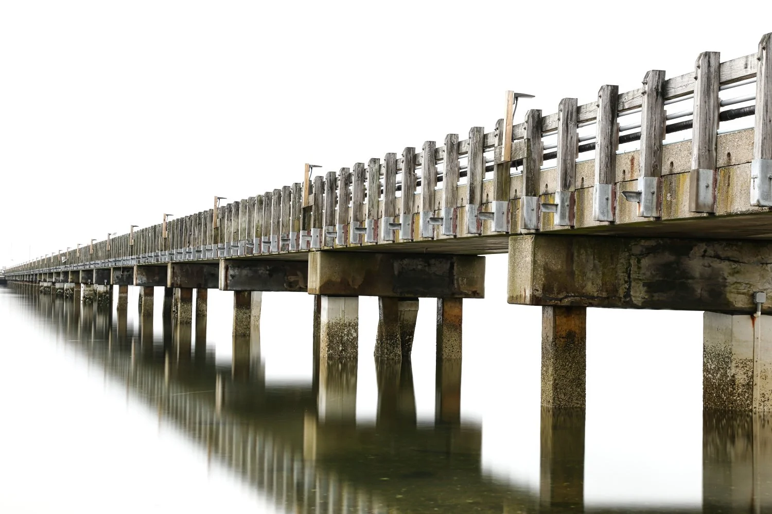 Long wooden pier extending over water, supported by concrete pillars, with a simple wooden railing along the sides.