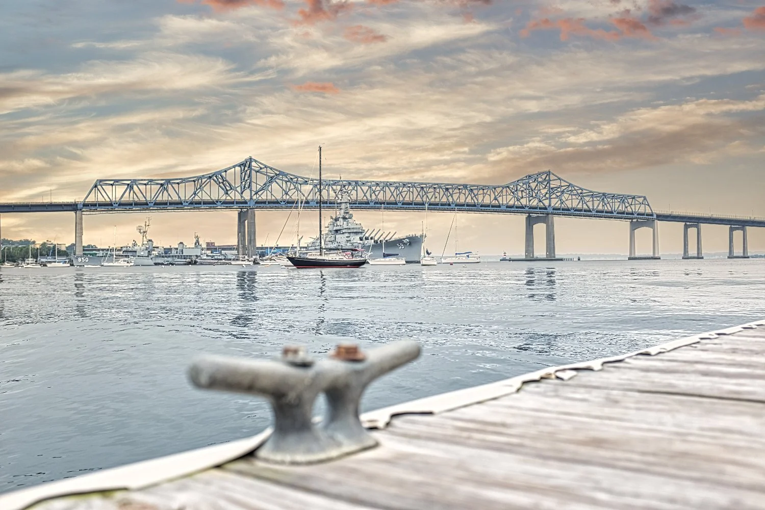 A view of a bridge over water with several boats and ships docked below during sunset or sunrise, with a weathered dock or pier in the foreground.