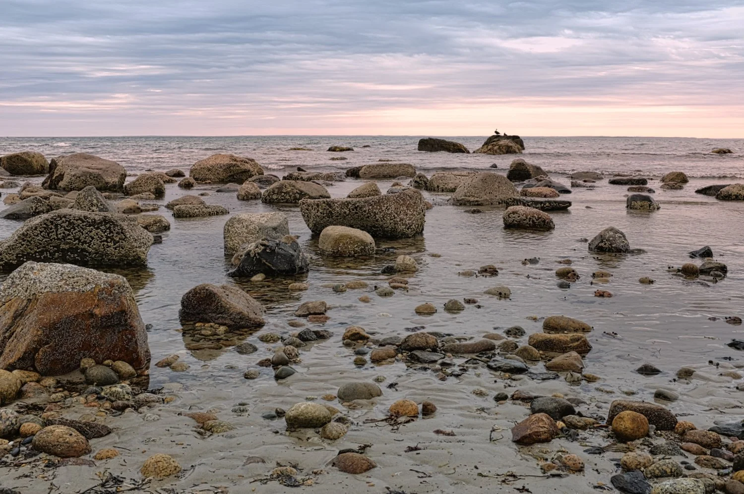 Horizon view of a rocky beach with calm water, large rocks along the shoreline, and a cloudy sky at sunset or sunrise.