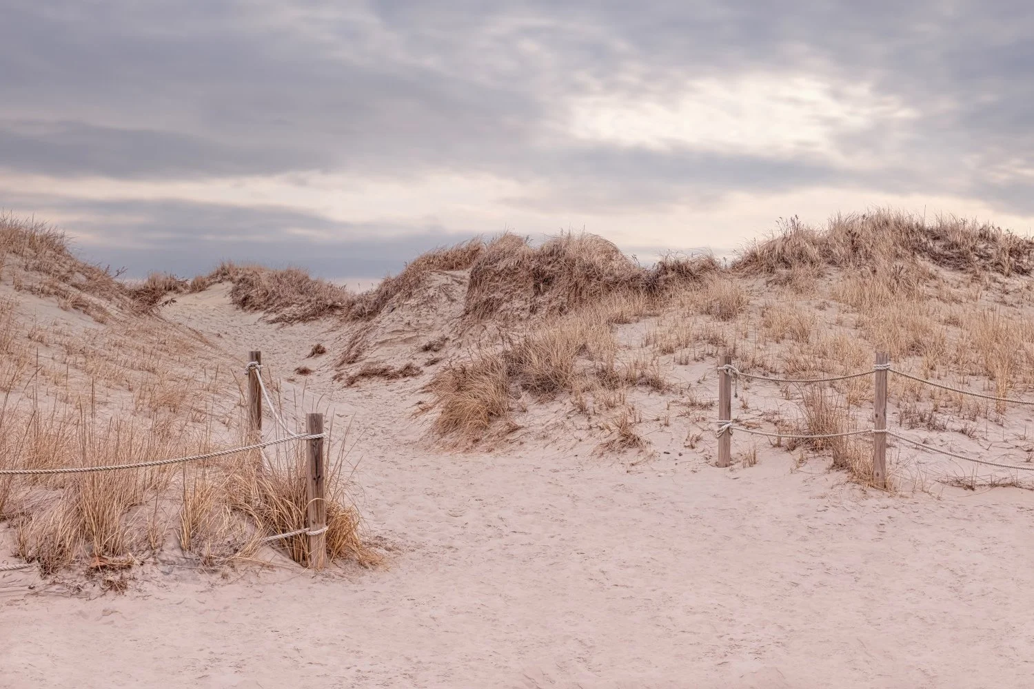 A sandy pathway lined with wooden posts and ropes leads through sand dunes covered with dry grass, under a cloudy sky.