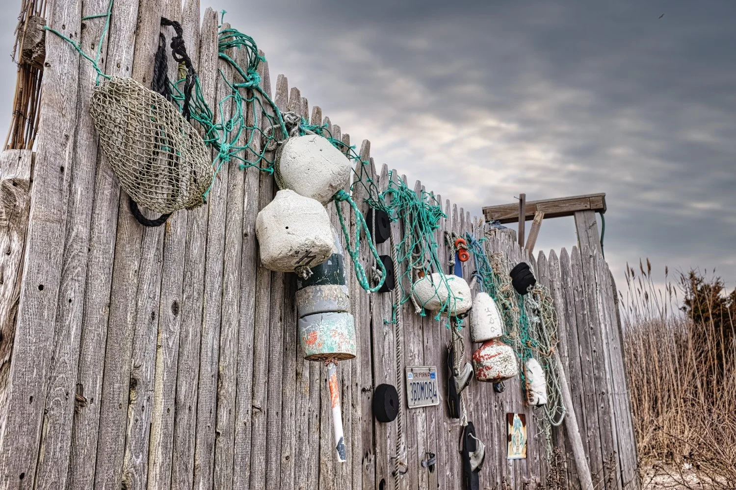 A weathered wooden fence decorated with various buoys, ropes, and fishing gear hanging from it on a cloudy day.