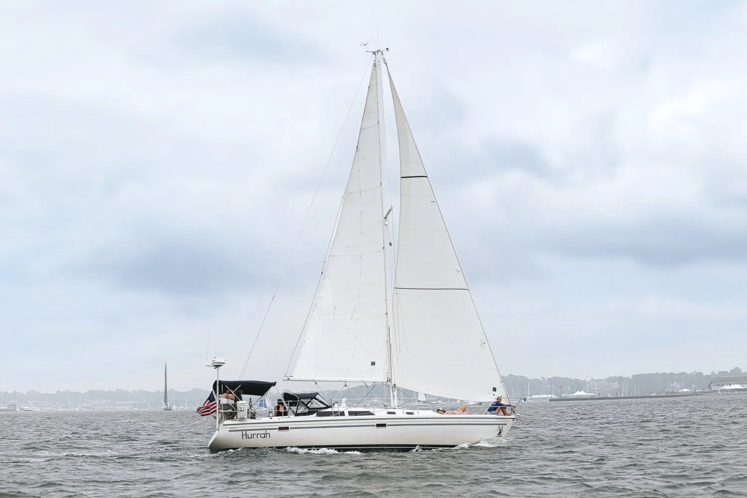 A sailboat named Hurrah with its sails up, sailing on a body of water under cloudy skies, with a distant shoreline visible in the background.