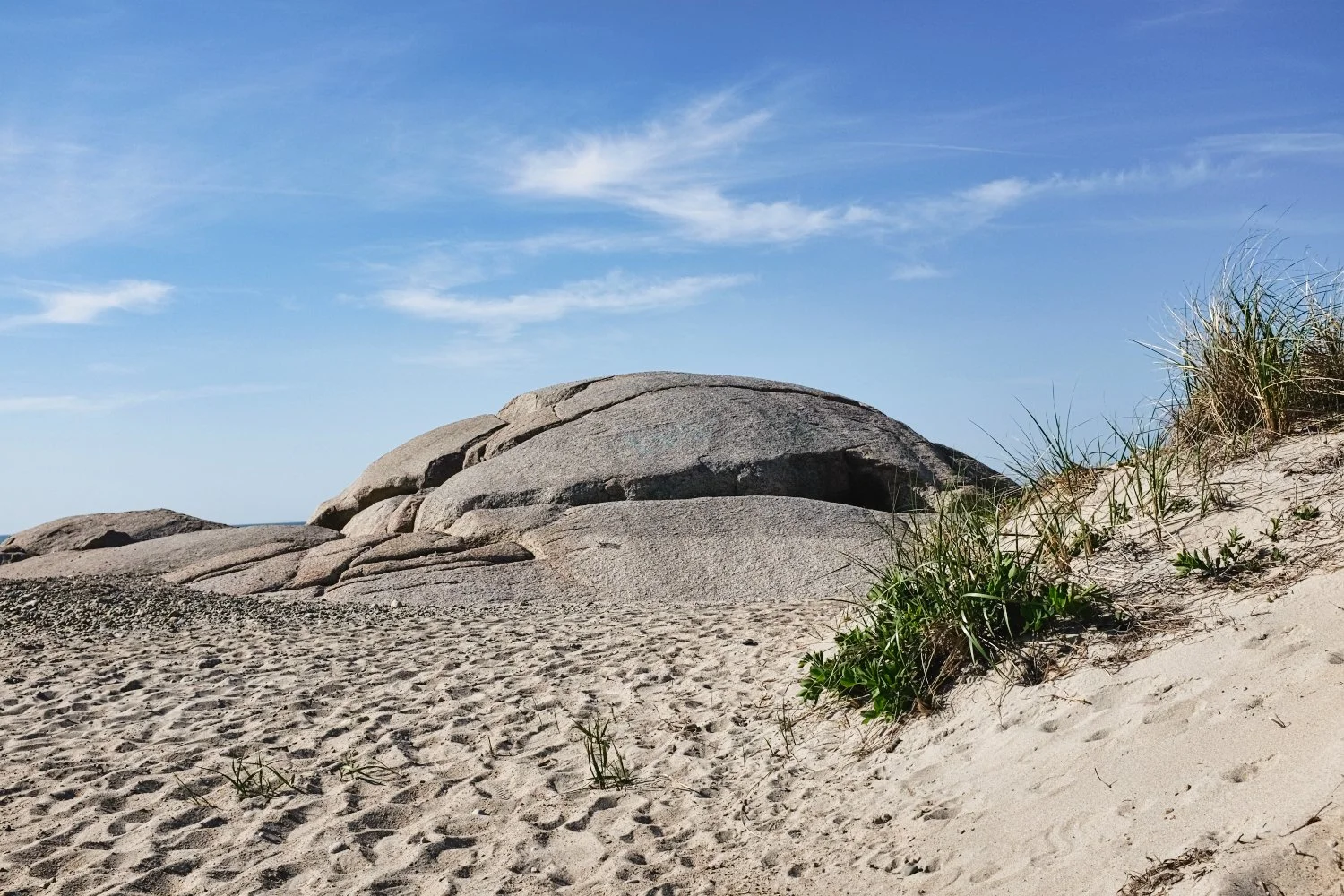 Sandy beach with large rocks and dune grass, under a blue sky with a few wispy clouds.