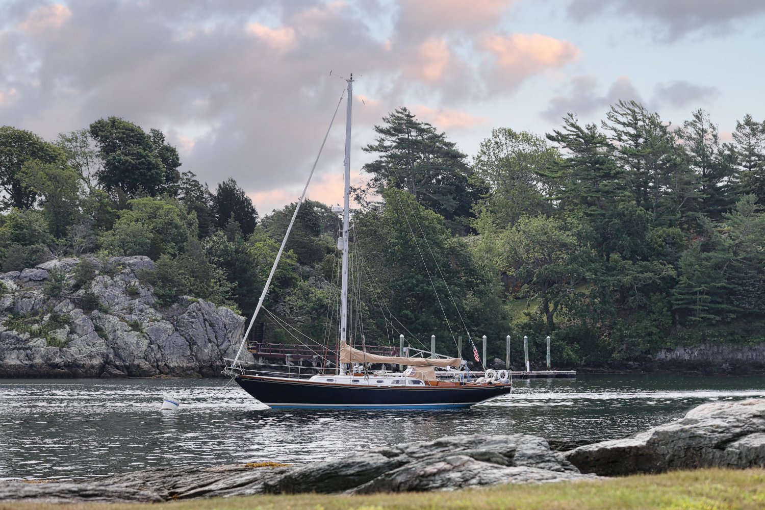 A sailboat sailing in a body of water near a rocky shoreline with lush green trees and a cloudy sky in the background.