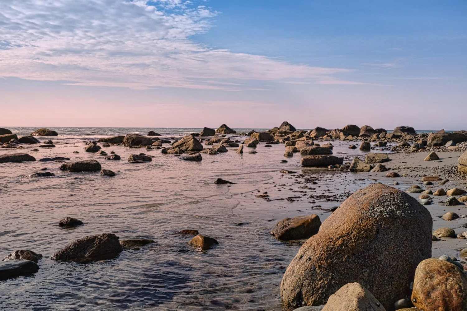 A rocky beach with large rocks scattered along the shore, calm water, and a partly cloudy sky during sunset or sunrise.