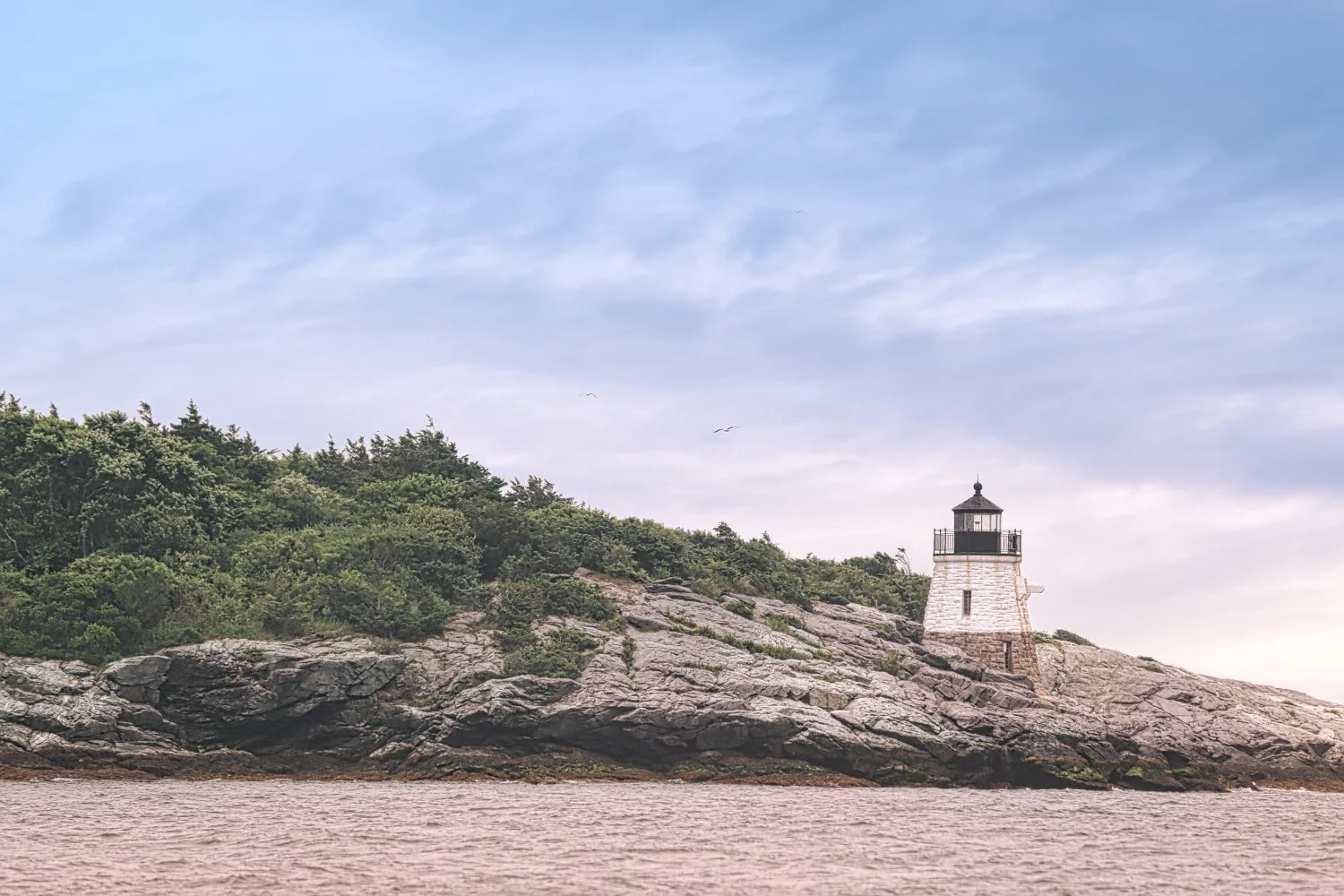 Lighthouse on rocky coastal area with trees and a cloudy sky.