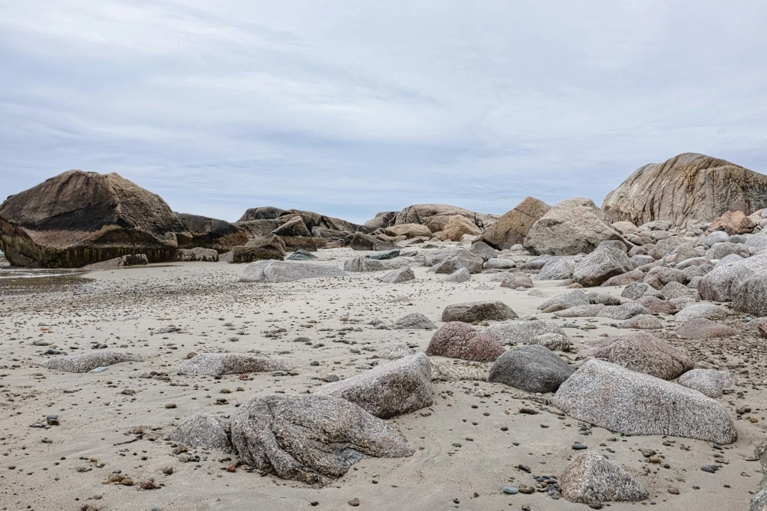 Rocky beach with large and small stones scattered on sandy shore under cloudy sky.