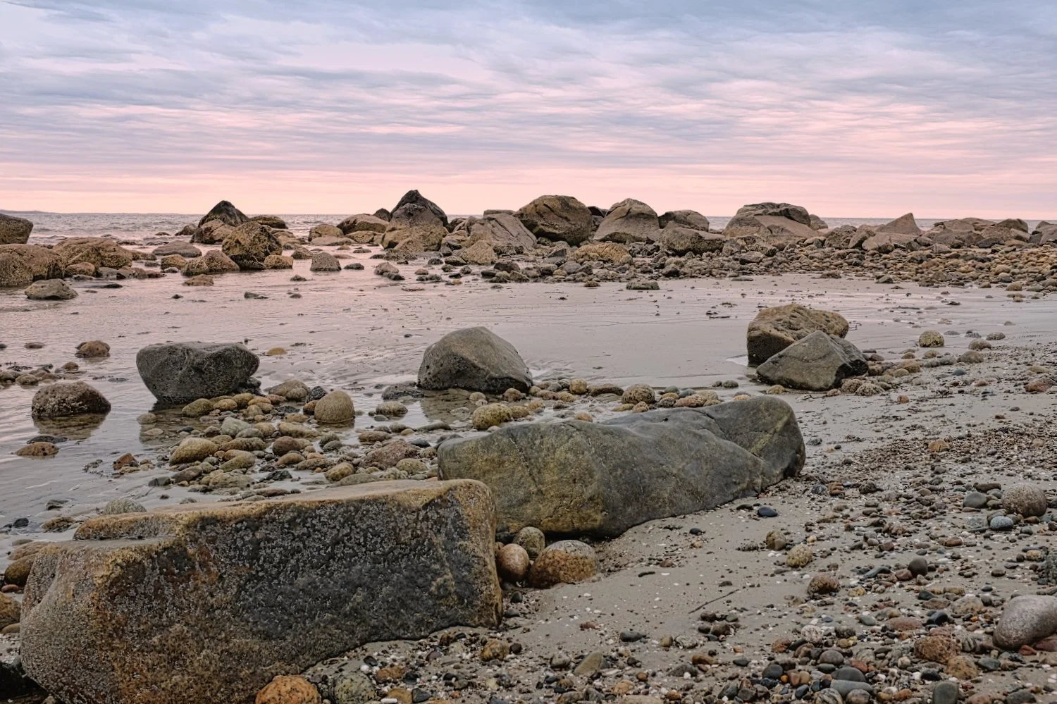 A rocky beach at sunset with large rocks scattered along the shore and a pastel-colored sky with pink and purple hues.