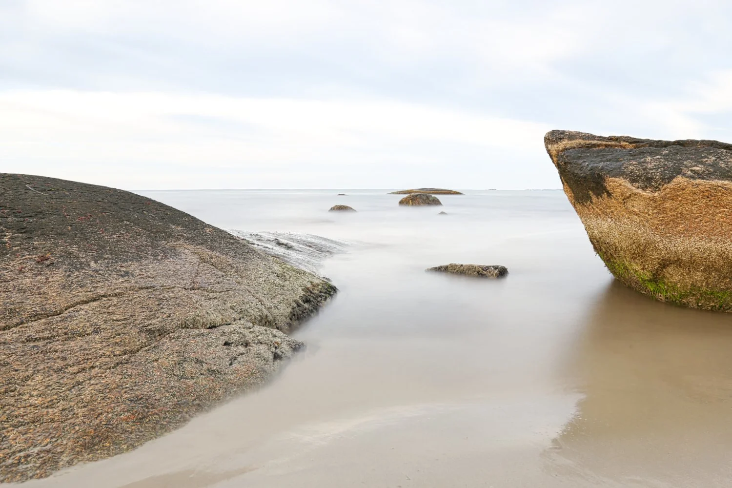 Calm coastal scene with large rocks partially submerged in water, a sandy beach, and a cloudy sky in the distance.