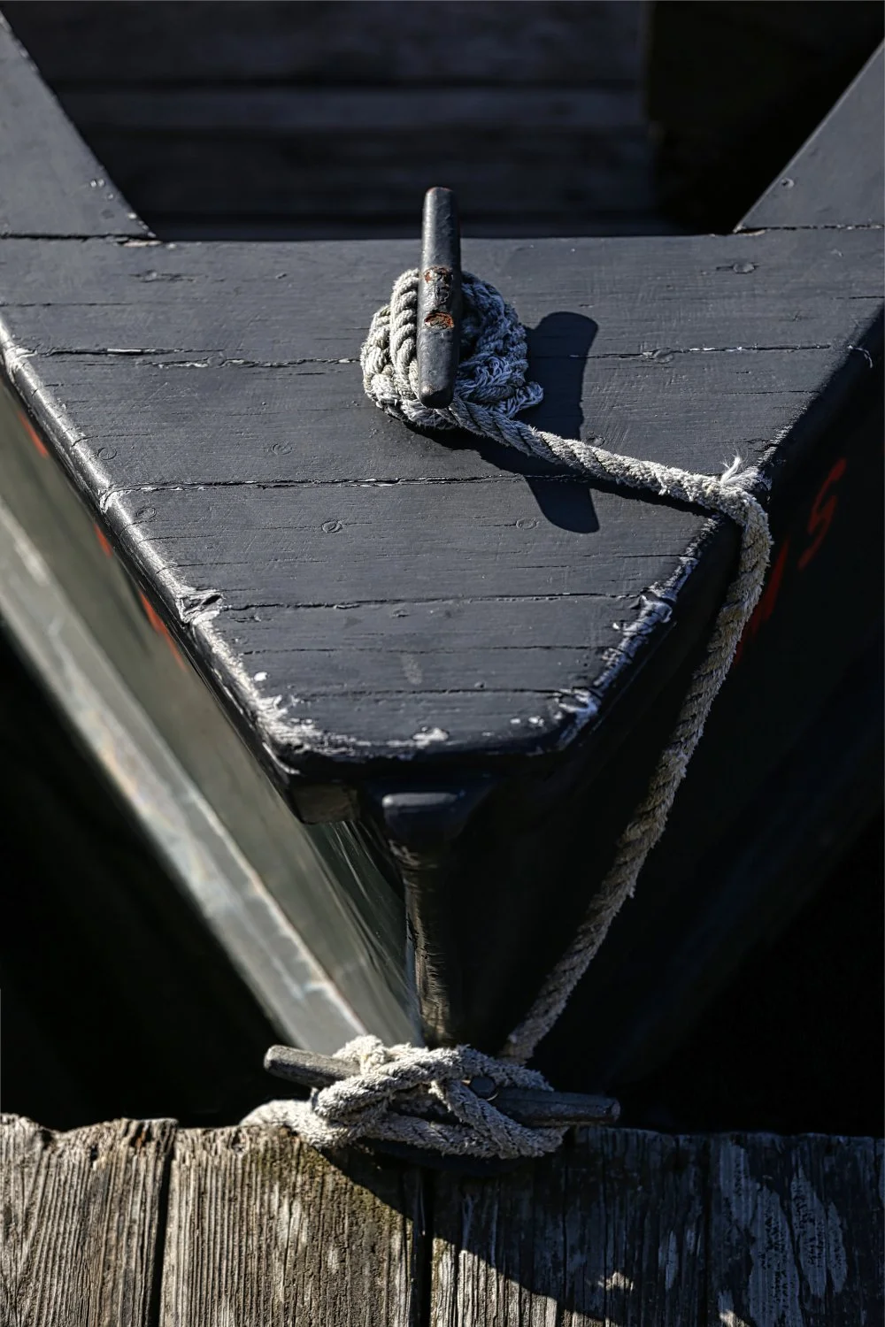 A close-up of a cleat with a coiled rope on it, attached to a wooden dock or boat platform, with weathered black paint and visible wood grain.
