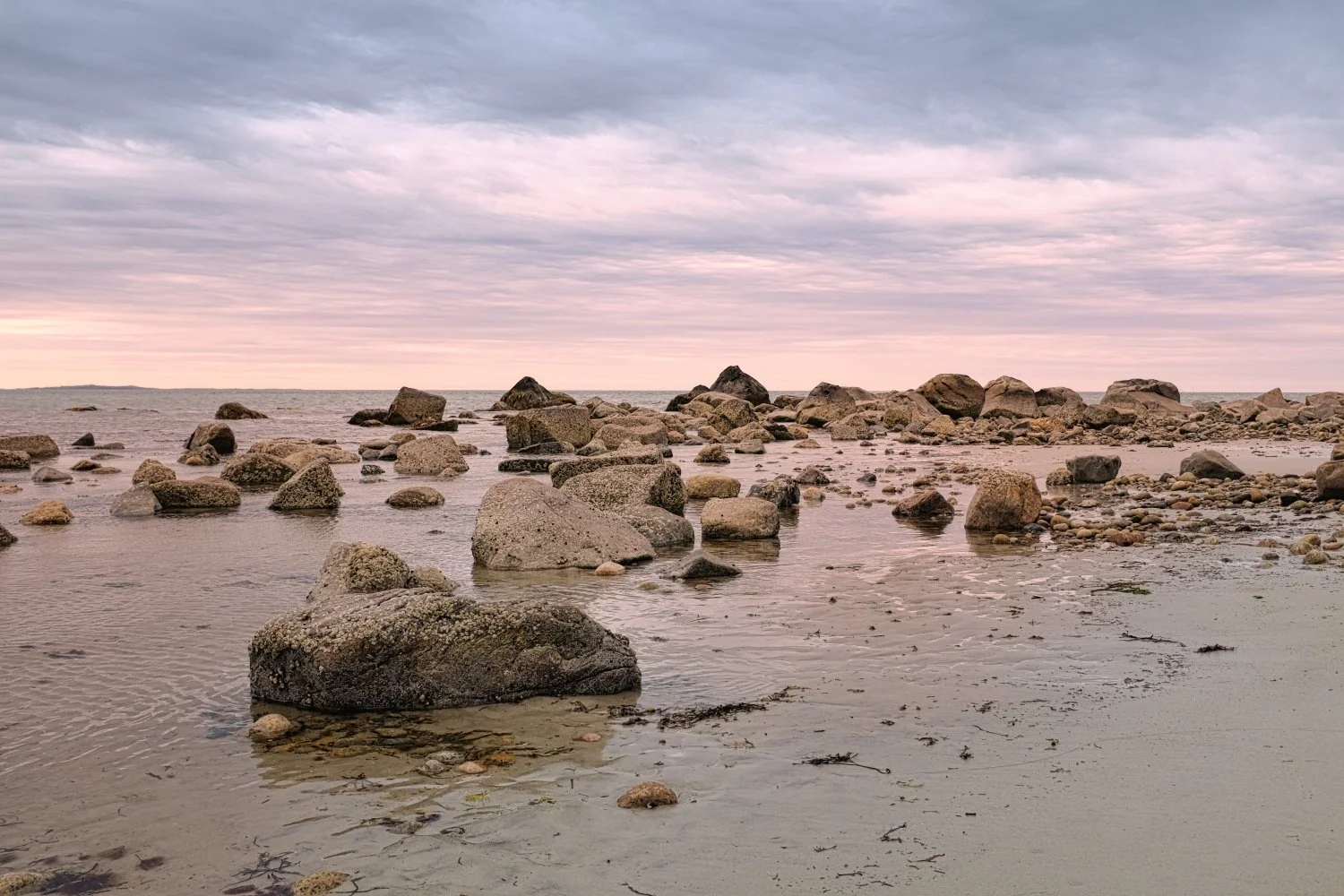 A rocky shoreline at sunset with scattered boulders and a cloudy sky.