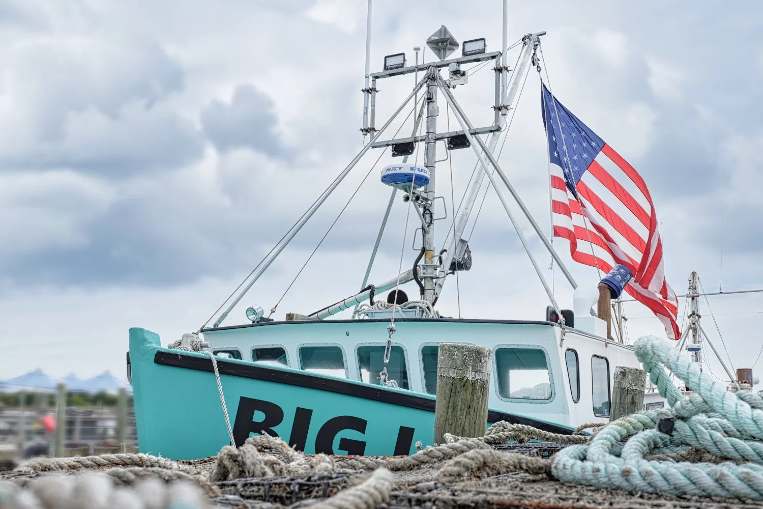 A boat docked at a pier with an American flag flying at the stern. The boat has the name "BIG" visible on the hull and is equipped with various antennas and navigation equipment. Ropes and wooden posts are in the foreground, and a cloudy sky is in th