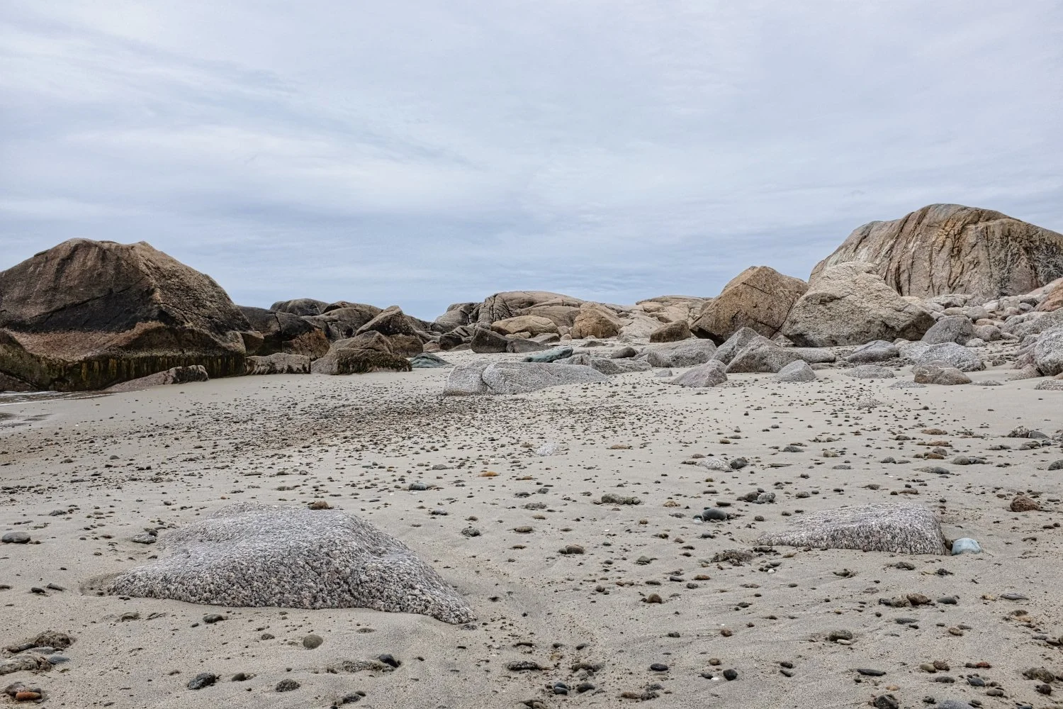 A beach scene with large rocks and sandy shore, cloudy sky overhead.
