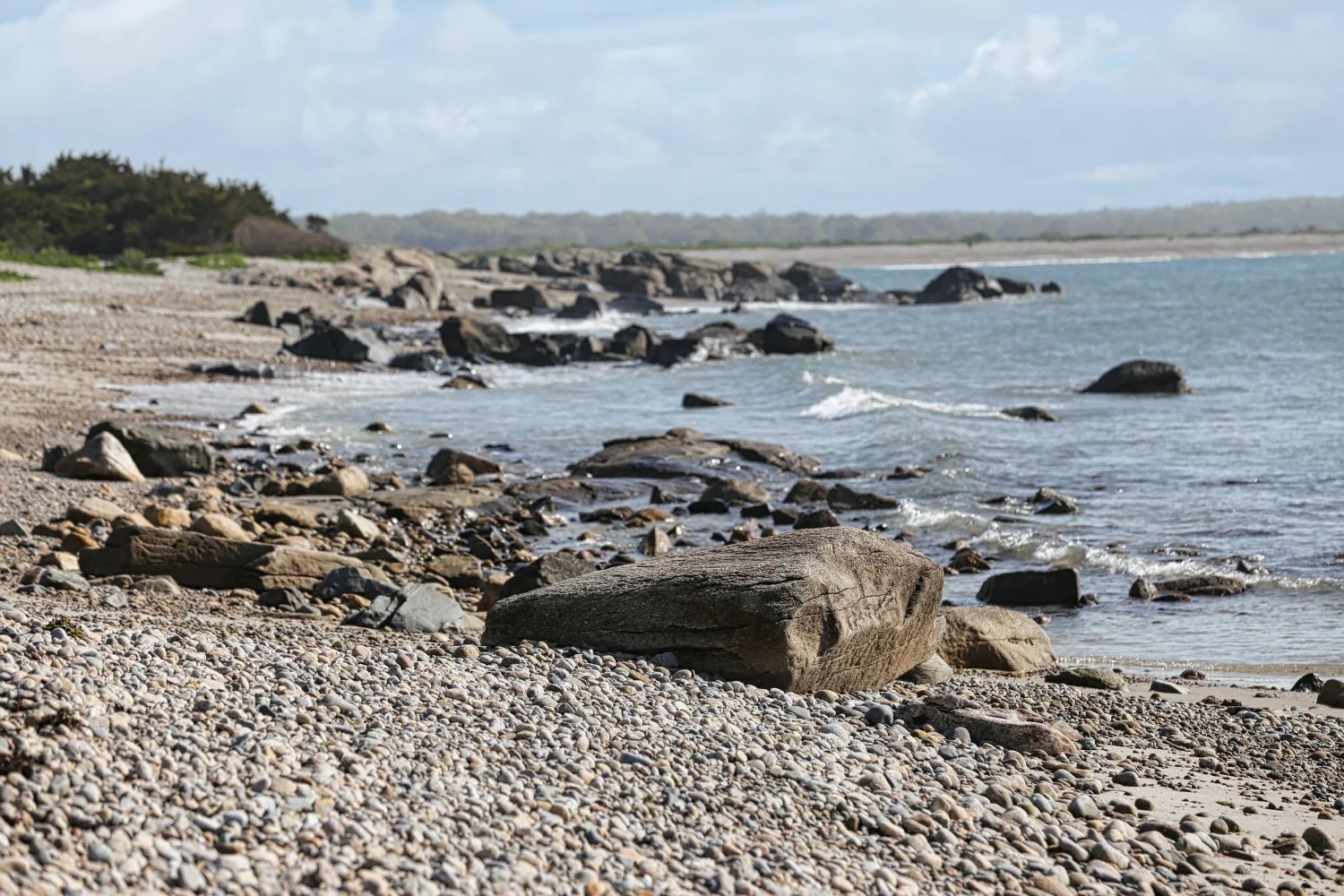 A rocky beach with large stones and pebbles along the shoreline, ocean waves, and a cloudy sky.